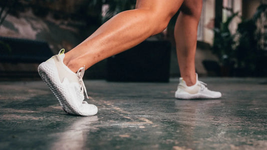 Woman stretching indoors wearing Vivobarefoot Motus Strength shoes in bright white, showcasing barefoot flexibility and grip on gym flooring